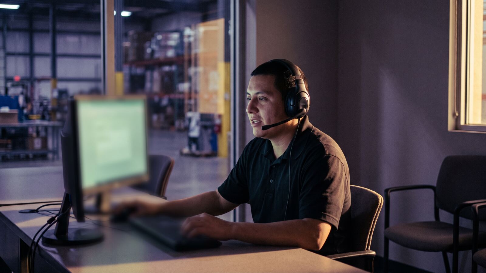 A new employee at a desk in a training room wearing a headset, speaking aloud to a screen, a warehouse floor visible through glass behind.
